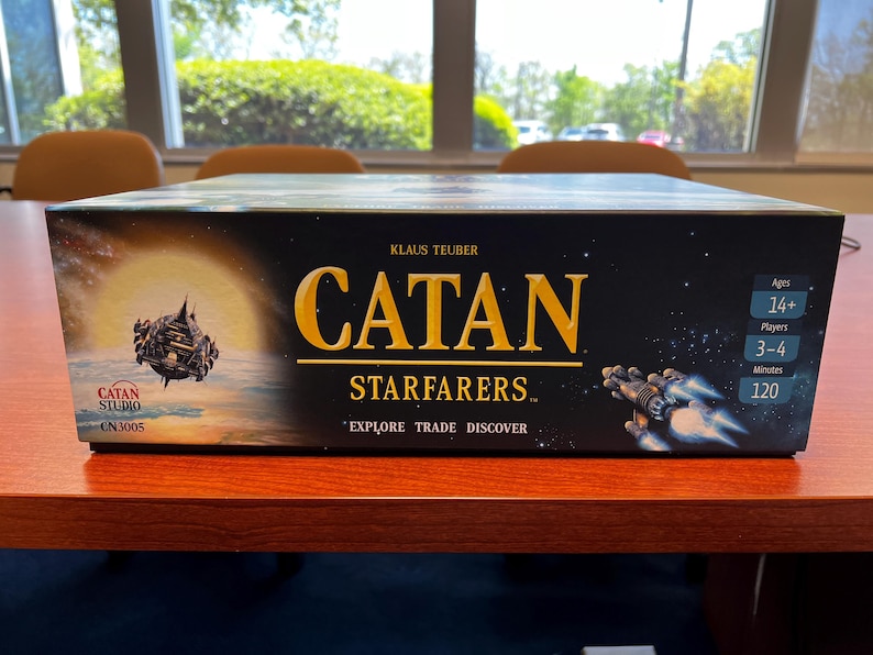 May include: A black box game with the title "CATAN STARFARERS" in gold lettering. The game is for 3-4 players, ages 14 and up, and takes 120 minutes to play.