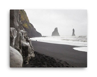May include: A black sand beach in Iceland with a rocky cliff on the left and two tall rock formations in the distance. The water is a light blue and the sky is grey.
