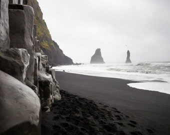 May include: A black sand beach with large rock formations in the distance. The beach is bordered by a rocky cliff on the left side of the image. The waves are crashing on the shore.