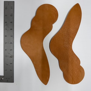 Puede incluir: Dos piezas de cuero marrón con forma de pies, probablemente para manualidades o trabajo en cuero.