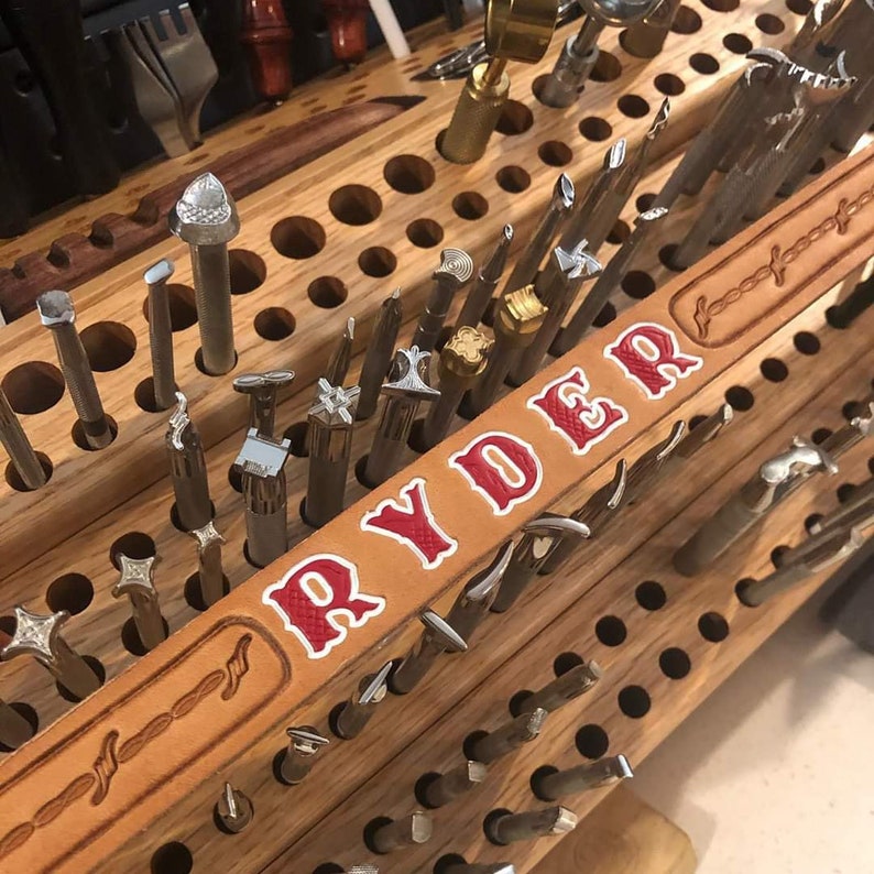 K&ouml;nnte beinhalten: Ein Holzwerkzeughalter mit einem Lederg&uuml;rtel, auf dem der Name "RYDER" in roten Buchstaben gepr&auml;gt ist. Der G&uuml;rtel ist mit einem geflochtenen Muster verziert. Der Halter ist mit verschiedenen Lederbearbeitungswerkzeugen gef&uuml;llt.