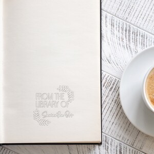 May include: A white notebook with a black spine and the text "From the Library of Samantha Ott" in a grey wreath design. The notebook is on a white wooden surface with a white saucer and a cup of coffee.