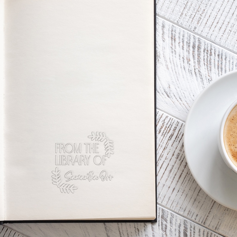 May include: A white notebook with a black spine and the text "From the Library of Samantha Ott" in a grey wreath design. The notebook is on a white wooden surface with a white saucer and a cup of coffee.
