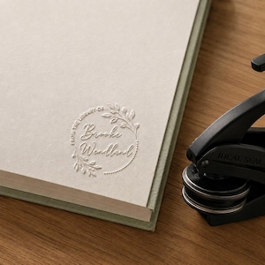 May include: A black metal embosser tool positioned next to a cream-colored book page. The page features a circular embossed design with the text "FROM THE LIBRARY OF" and "Brooke Wendland" surrounded by a delicate leaf and vine wreath pattern.