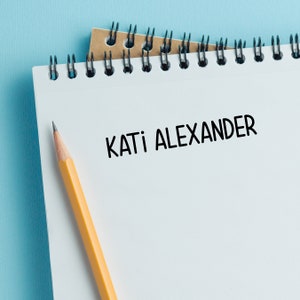 May include: A white spiral notebook with the name "KATI ALEXANDER" written in black ink. A yellow pencil is lying on the notebook.