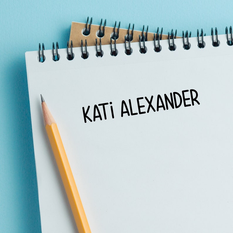 May include: A white spiral notebook with the name "KATI ALEXANDER" written in black ink. A yellow pencil is lying on the notebook.