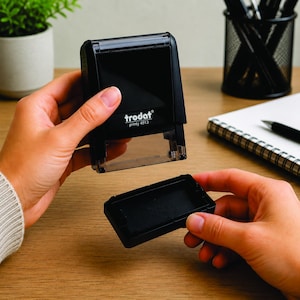 May include: A black Trodat printy 4913 self-inking stamp with a clear base, held over its ink pad. The stamp is on a wooden surface, with a plant, pen holder, and notepad in the background.