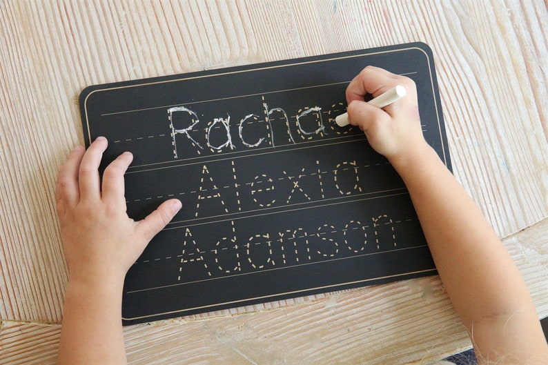 Puede incluir: Una pizarra negra con ribete dorado y l&iacute;neas blancas. Un ni&ntilde;o est&aacute; trazando los nombres "Rachael", "Alexa" y "Adamson" con un marcador blanco.
