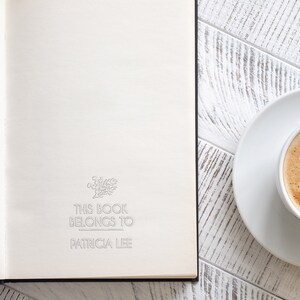 May include: A white book with the text "This book belongs to Patricia Lee" on the front cover. A white saucer with a cup of coffee is on a white wooden table next to the book.