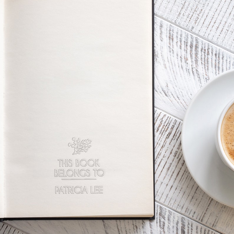 May include: A white book with the text "This book belongs to Patricia Lee" on the front cover. A white saucer with a cup of coffee is on a white wooden table next to the book.