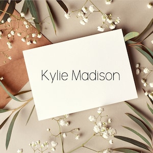 May include: A white card with the name "Kylie Madison" written in black text. The card is surrounded by small white flowers and green leaves on a light brown background.