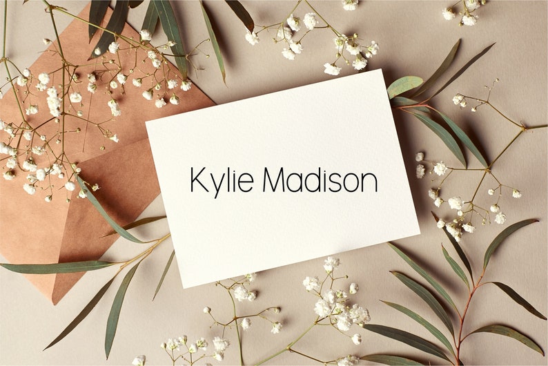 May include: A white card with the name "Kylie Madison" written in black text. The card is surrounded by small white flowers and green leaves on a light brown background.
