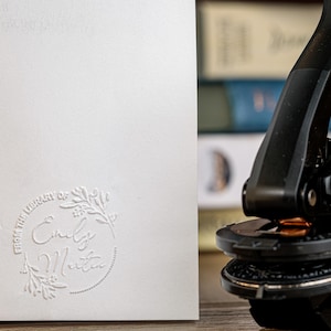 May include: A white card with embossed text reading "FROM THE LIBRARY OF Emily Martin" within a circular floral design. A black seal press is in the background, suggesting stationery or book-related items.