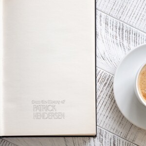 May include: A white book with the text "from the library of PATRICK HENDERSEN" on the front cover. A white saucer with a cup of coffee is on a white wooden table next to the book.