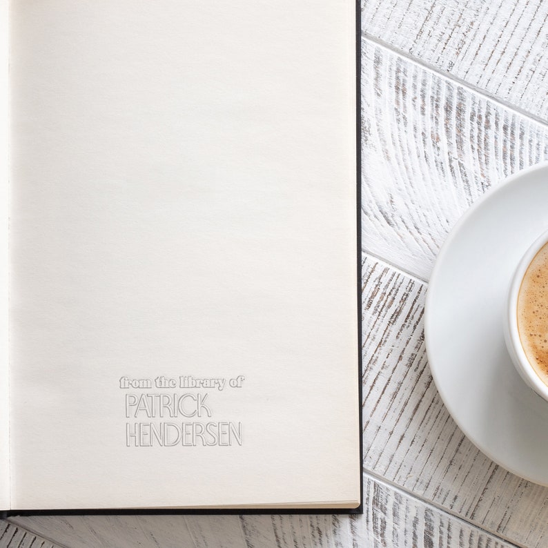 May include: A white book with the text "from the library of PATRICK HENDERSEN" on the front cover. A white saucer with a cup of coffee is on a white wooden table next to the book.