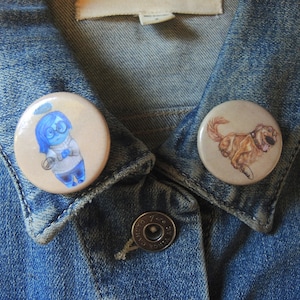 May include: Two button pins on a denim jacket. One pin features a cartoon character with blue hair and a blue dress, the other pin features a cartoon dog.