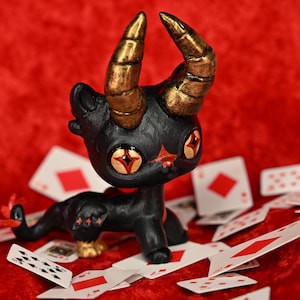 May include: A black and gold demon figurine with red eyes, sitting on a red surface with playing cards scattered around it.