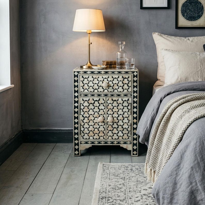 A rectangular bedside table featuring a black and white geometric inlay pattern. The piece includes one top drawer and two lower cabinet doors, each fitted with small, round, light-coloured knobs. The frame is finished with a dark border.