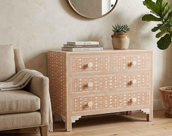 May include: A rectangular wooden dresser featuring three drawers with a light peach and white geometric inlay pattern. Each drawer is equipped with two round white knobs. The dresser stands on four short, stepped feet and has a smooth finish.