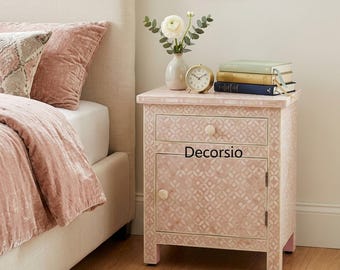 May include: A pink bedside table with a geometric pattern, a drawer, and a cabinet door. A small gold clock, a white vase with flowers, and a stack of books sit on top. The text "Decorsio" is visible on the front.