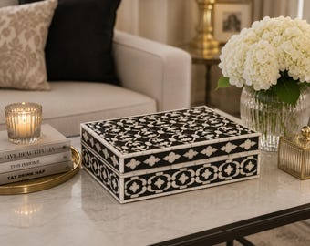 May include: A decorative black and white box with a geometric pattern sits on a marble-topped table. The box is rectangular and has a hinged lid. A lit candle and books are on a gold tray.