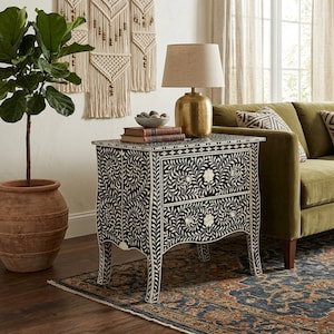 Könnte beinhalten: Ein Nachttisch mit zwei Schubladen und einem schwarz-weißen Blumenmuster. Eine goldene Lampe, Bücher und eine Schale stehen darauf. Der Raum umfasst ein grünes Sofa und einen gemusterten Teppich. Wohnzimmer-Atmosphäre.