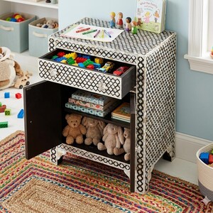 Könnte beinhalten: Ein schwarz-weiß gemusterter Nachttisch mit einer offenen Schublade voller Spielzeug. Der Schrank hat eine offene Tür darunter, die Regale mit Büchern und Stofftieren freigibt. Ein bunter Teppich liegt davor.