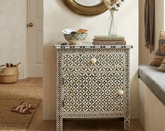 May include: A black and white patterned bedside table with a drawer and a cabinet door. The table has a decorative floral design and round knobs. A decorative bowl, books, and a vase with dried plants sit on top of the table.