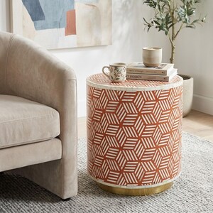 Può includere: Un tavolino cilindrico caratterizzato da un motivo geometrico a cubi ripetuti in arancione bruciato e bianco, rifinito con una base circolare color oro. Il piano ospita una tazza in ceramica con stampa floreale e due libri con una candela.