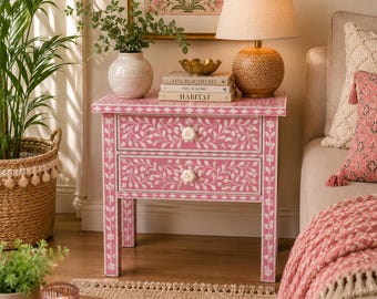 May include: A pink bedside table with two drawers, adorned with a white floral inlay design. The table features white flower-shaped drawer pulls. A lamp, books, and a vase with greenery sit atop the table. The table is next to a beige sofa with pink pillows.