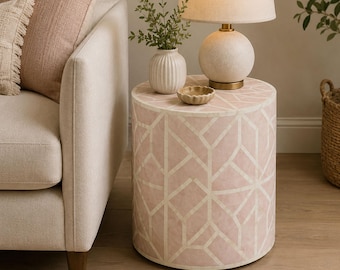May include: A cylindrical side table featuring a soft pink finish with an inlaid white geometric pattern. The table surface holds a small, round, textured beige lamp with a cream shade, a small white ribbed vase with greenery, and a small bowl.