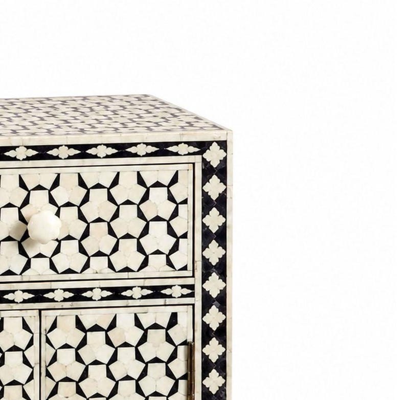 A rectangular bedside table featuring a black and white geometric inlay pattern. The piece includes one top drawer and two lower cabinet doors, each fitted with a small round white knob. The frame is decorated with a repeating floral border.