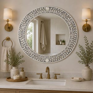 May include: A round wall mirror featuring a dark gray frame inlaid with a white mother-of-pearl leaf pattern and floral accents. The mirror is centered above a bathroom vanity with a white sink, brass faucet, and two matching wall sconces.