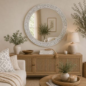 May include: A round wall mirror with a light gray frame featuring a white floral and leaf pattern. The mirror is mounted on a wall above a wooden console table with cane cabinet doors, reflecting a room with a lamp and a framed picture.