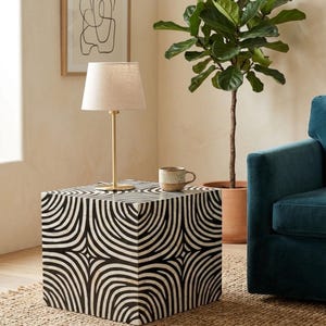 May include: A square side table featuring a black and white geometric pattern of curved lines. On top sits a table lamp with a thin gold-toned metal base and a light beige fabric shade, alongside a speckled ceramic mug with a handle.