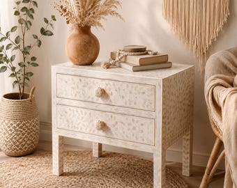May include: A cream-colored nightstand with two drawers, adorned with a floral pattern. The nightstand has decorative knobs and is placed on a jute rug. A vase with dried plants and books sit on top.