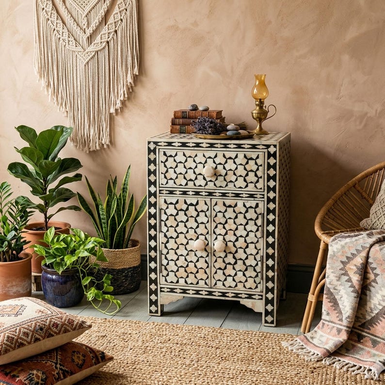 A warm, boho-chic room setting featuring a black and white geometric bone inlay nightstand styled with antique books, a brass oil lamp, and surrounding potted indoor plants next to a rattan chair.