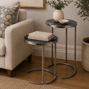 Op de afbeelding: Een set van twee bijzettafels met ronde zwarte marmeren bladen en zilverkleurige metalen frames met een gehamerde textuur. De tafels hebben gebogen, open onderstellen. Op de kleinere tafel liggen twee boeken met de titels Live Beautifully en This Is Home.