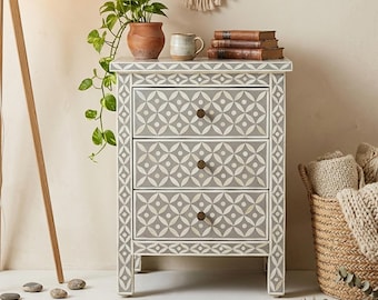 May include: A gray three-drawer nightstand with a white geometric pattern. The nightstand has bronze knobs and is placed on a patterned rug. A potted plant, mug, and books are on top. A woven basket and a floor lamp are also in the image.