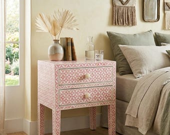 May include: A two-drawer bedside table with a pink and white geometric pattern. The table has cream-coloured knobs and matching patterned legs. It is decorated with a vase, books, and a glass. The room has a neutral colour palette.