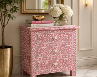 May include: A pink three-drawer nightstand with a white floral inlay design. The nightstand has white flower-shaped drawer pulls. A gold bowl and books are on top of the nightstand. A gold-colored plant pot is on the left.