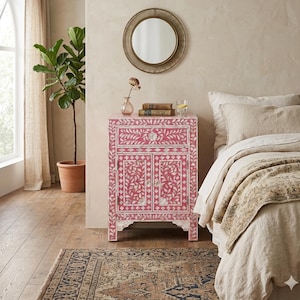 Puede incluir: Una mesita de noche rosa con un diseño de incrustaciones florales. El armario tiene un cajón y dos puertas, y está decorado con motivos florales blancos. Un espejo redondo cuelga sobre el armario, y una planta en una maceta de terracota está a la izquierda.