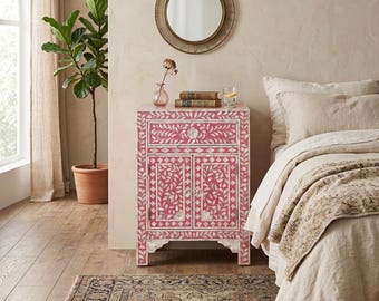 May include: A pink bedside cabinet with a floral inlay design. The cabinet has a drawer and two doors, and is decorated with white floral patterns. A round mirror hangs above the cabinet, and a plant in a terracotta pot is to the left.