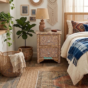 May include: A bedroom interior with a patterned nightstand, a woven lamp, and a bed with a blue throw. A large potted plant and a woven basket enhance the room's decor. The nightstand has two drawers with silver knobs.