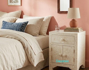May include: A bedroom scene featuring a bed with beige bedding and decorative pillows. A white nightstand with floral inlay and a lamp with a terracotta base sit beside the bed. A framed mirror hangs on the pink wall.