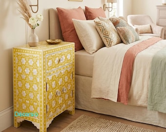 May include: A yellow and white floral patterned nightstand with two drawers and a cabinet door. The nightstand is next to a bed with pillows and a blanket. A beige rug is on the floor.