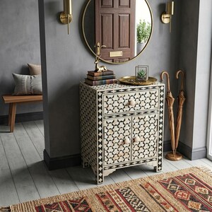 A rectangular cabinet featuring a black and white geometric inlay pattern, one top drawer, and two lower doors with round wooden knobs. The cabinet is constructed with a frame-like border and stands on four short, straight legs.