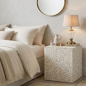 Peut inclure: Une table de chevet carrée avec un motif incrusté de feuilles blanches et beiges. Sur le dessus se trouvent une lampe à base en laiton avec un abat-jour beige, un pichet en céramique blanche et un verre transparent près d'un lit aux draps beiges.