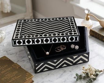 May include: A black and white decorative jewelry box with a geometric chevron pattern on the lid and base. The open box reveals a black velvet interior with a pearl necklace, rings, and earrings. The box is on a marble surface.