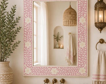 May include: A rectangular wall mirror featuring a wide frame with a pink background and an intricate white floral and vine pattern. The mirror is mounted above a stone sink with brass fixtures, reflecting a bathroom interior with a hanging light.